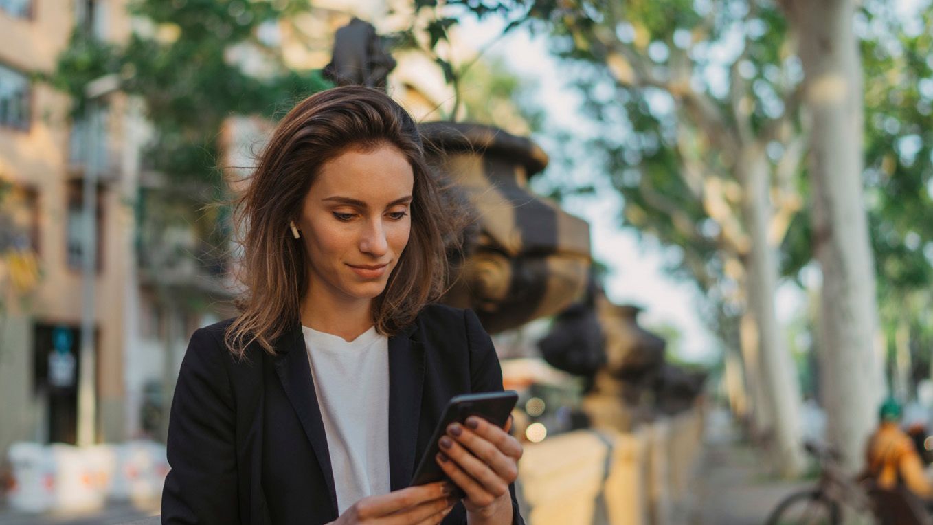 Ženska v poslovnem suknjiču stoji ob kamniti ograji v parku in gleda v pametni telefon.