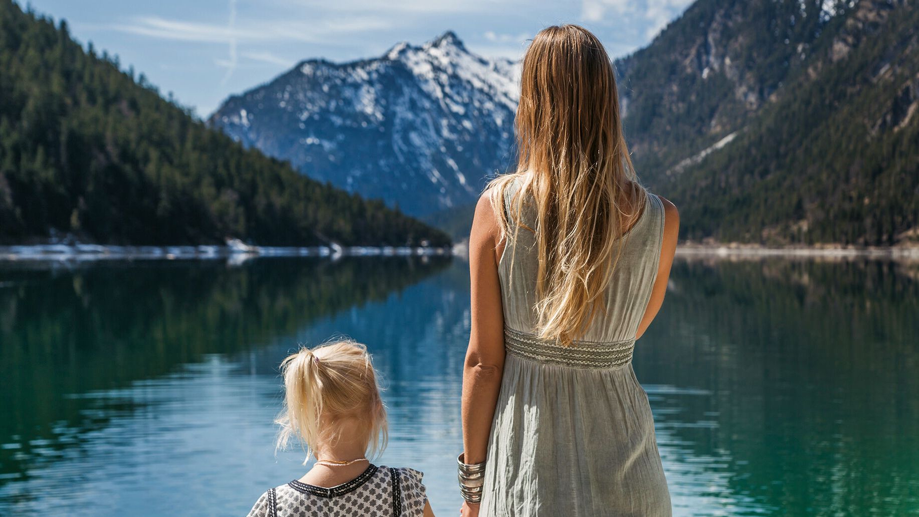 Mutter und Tochter stehen mit dem Rücken zur Kamera und betrachten vor Ihnen den Bergsee 