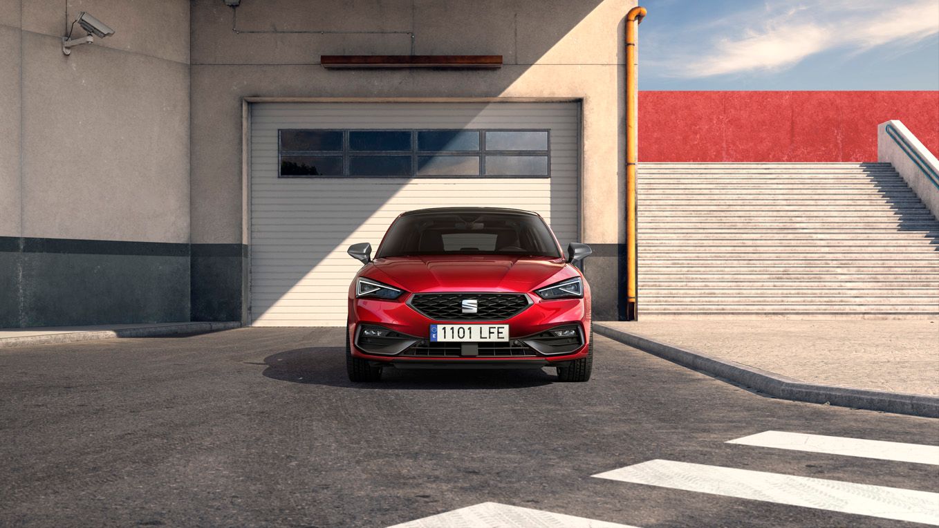 Rdeč SEAT Leon parkiran pred velikimi garažnimi vrati na dvorišču industrijske zgradbe.
