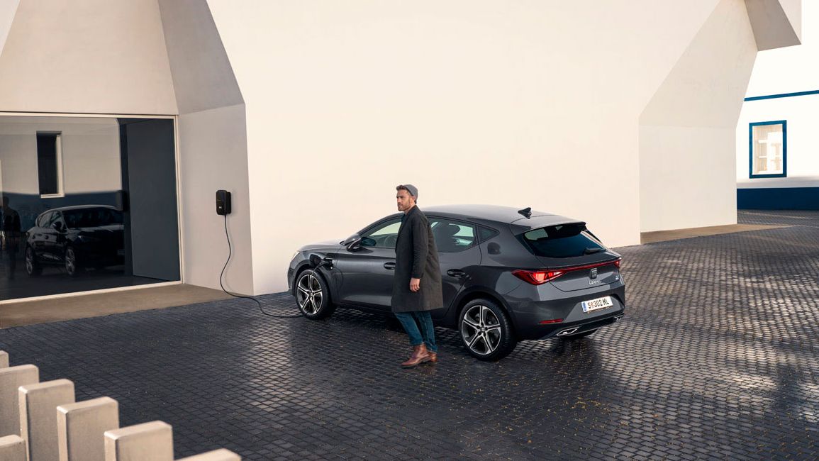 Der SEAT Leon Hybrid 5-Türer wird an einer Elektroladestation geladen während ein Mann davor steht und wartet 